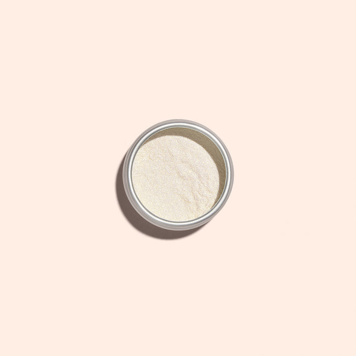 A small metal tin of Manucurist's Golden Glazed Effect Powder—fine, off-white powder that gives nails a pearly gold shimmer—sits centered on a light beige background, casting a soft left shadow.