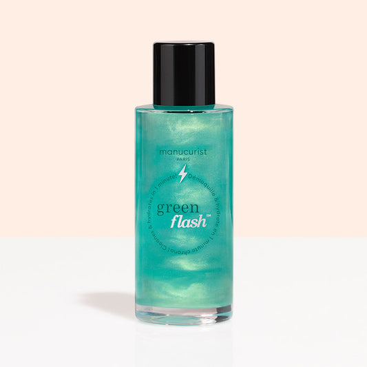 A clear bottle with a black cap, labeled “Manucurist Green Flash™ Gentle Glitter Remover,” holds shimmery teal, acetone-free liquid and is displayed against a light peach and white background.