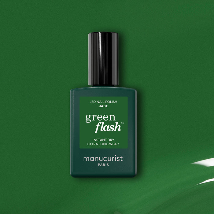 A bottle of Manucurist Jade Green Flash LED nail polish sits against a green backdrop, with a glossy green paint streak in the lower right corner.