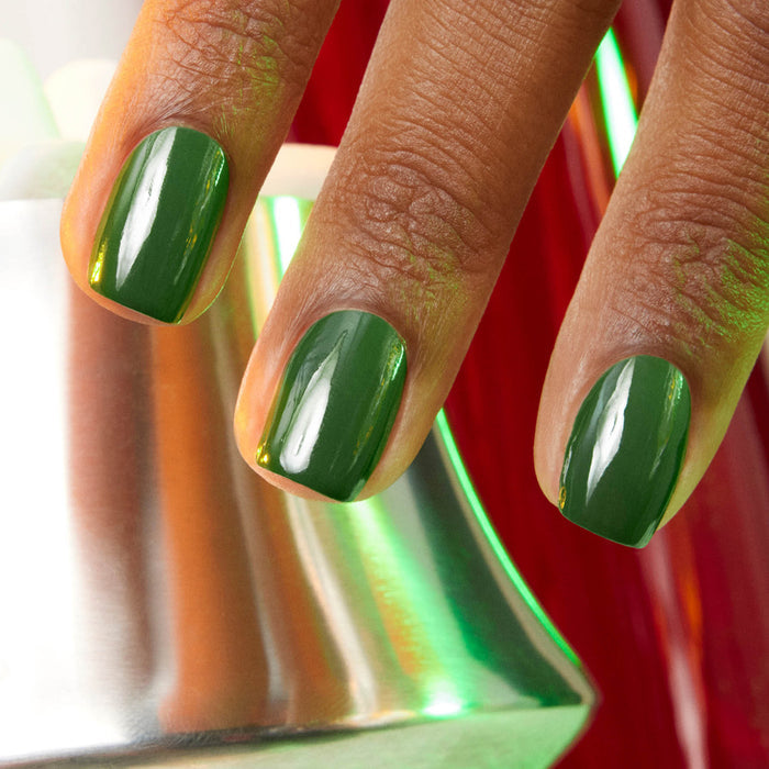 A close-up of three fingers with glossy Manucurist Jade LED gel polish, shown near a metallic surface reflecting green and red light.