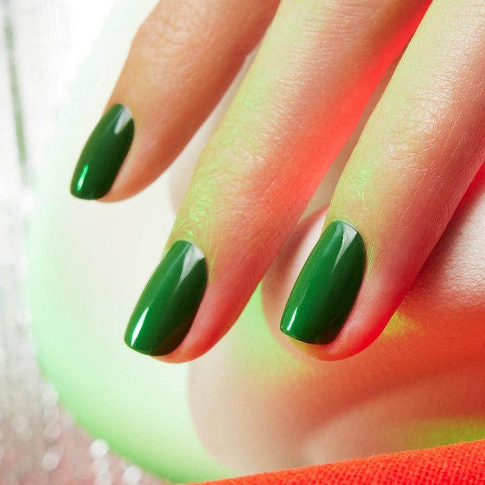A close-up of a hand with neatly manicured nails painted in Manucurist's glossy, vibrant Jade polish, set against a softly blurred background with touches of red and green light.