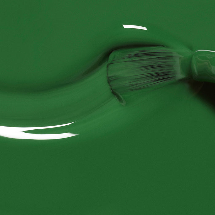 A close-up of a paintbrush applying Manucurist Jade, a glossy mineral green polish, across a smooth surface, with visible brush bristles and shiny reflections on the wet finish.