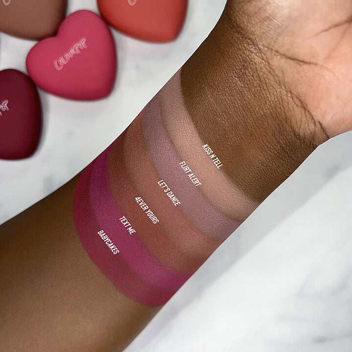 ColourPop Kiss 'N Tell Pressed Powder Blush swatched on deep skin, showing a soft matte rose‑beige payoff; additional blush swatches and pink heart‑shaped compacts appear blurred in the background on a marble surface.
