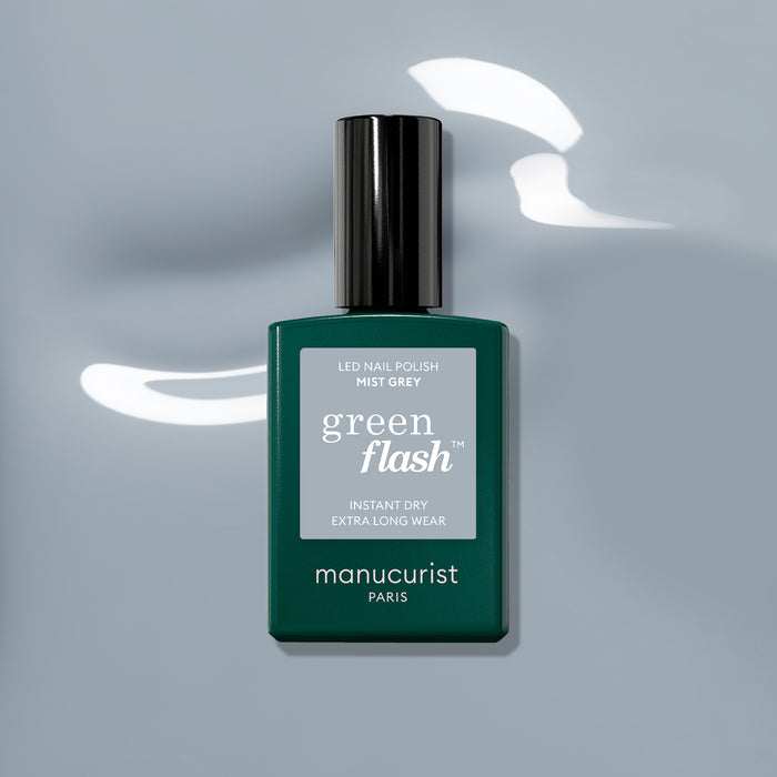 A bottle of Manucurist Mist Grey Green Flash LED gel polish sits on a glossy light grey surface, with reflections visible around its dark green rectangular base and black cap.