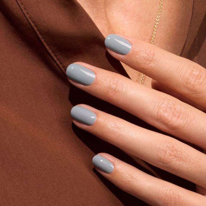 A close-up of a hand with neatly manicured nails painted in Manucurist's Mist Grey gel polish, resting on a brown fabric surface.