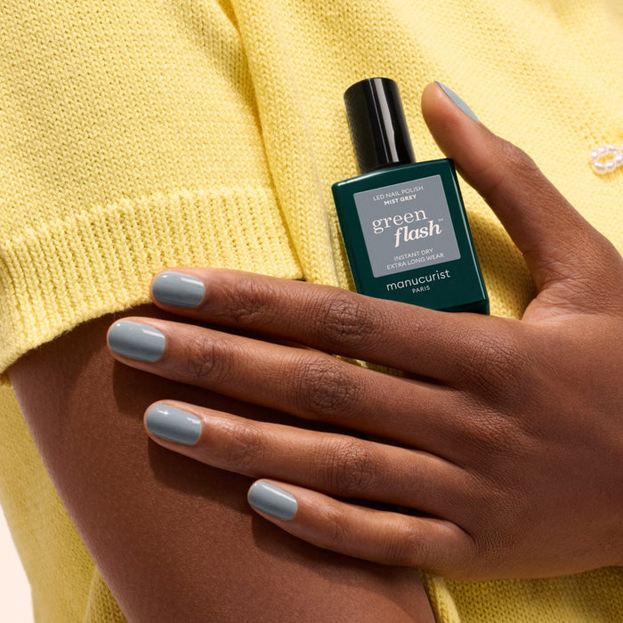 A person with medium brown skin displays their Mist Grey gel polish nails while holding a bottle of Manucurist nail polish against a yellow knit top.