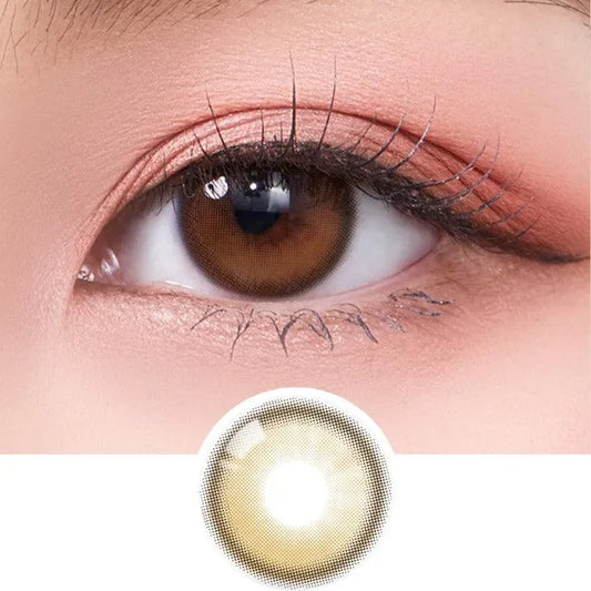A close-up of a brown eye wearing Olola Dahlia Brown by Olola, featuring the natural-looking brown non-prescription contact lens design below. The makeup is soft with peach-toned eyeshadow and defined eyelashes.