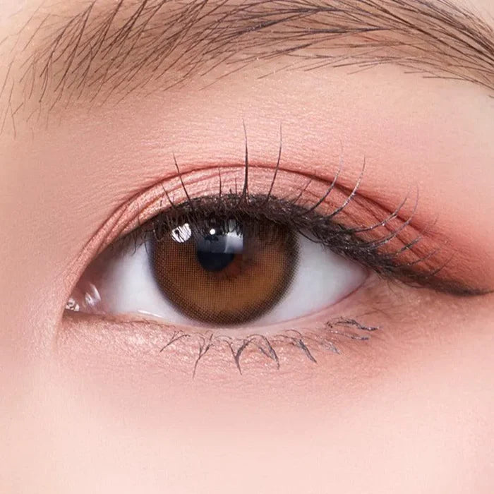 A close-up of an eye wearing Olola Dahlia Brown color contact lens by Olola, with long lashes and soft pink eyeshadow on smooth, flawless skin.