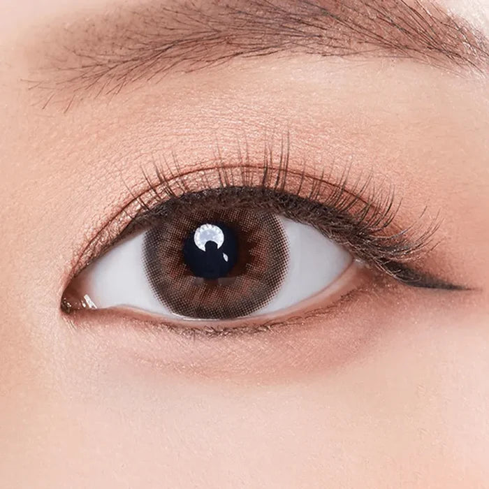 A close-up of a person's eye with natural makeup, long lashes, and Olola Dazzling 1-Day Rosie Hazel (10pk) contact lenses by Olola. The well-groomed brow, soft eyeshadow, and eyeliner beautifully enhance the look.