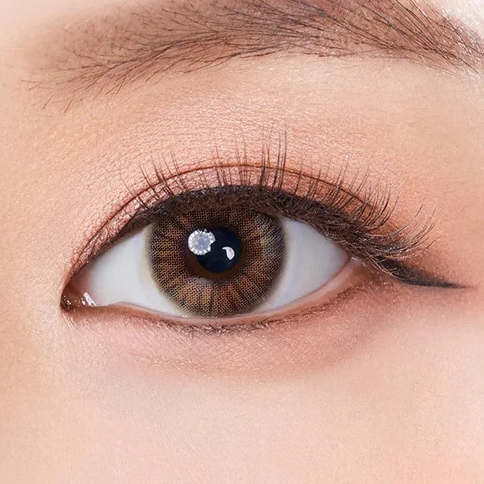 A close-up of an eye with a natural brown iris enhanced by Olola Heiress Brown contact lenses from Olola, long lashes, peach eyeshadow, a well-groomed brow, and subtle winged eyeliner.