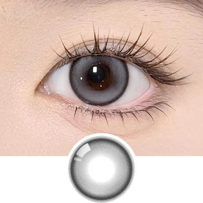 A close-up of an eye wearing Olola Meow Natural Coon Grey contacts from Olola, which enlarge the iris and feature long lashes. A single grey Olola lens is displayed separately below the eye.