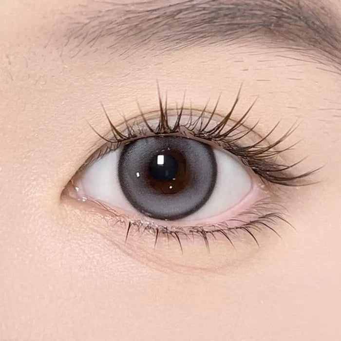 A close-up of a brown eye wearing Olola Meow Natural Coon Grey contact lenses by Olola, featuring long, curled eyelashes and smooth, light skin around the eye.