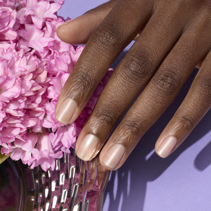 A hand with neatly manicured, Pearlescent by Manucurist nails in a natural nude with pink undertones rests next to pink flowers on a purple surface.