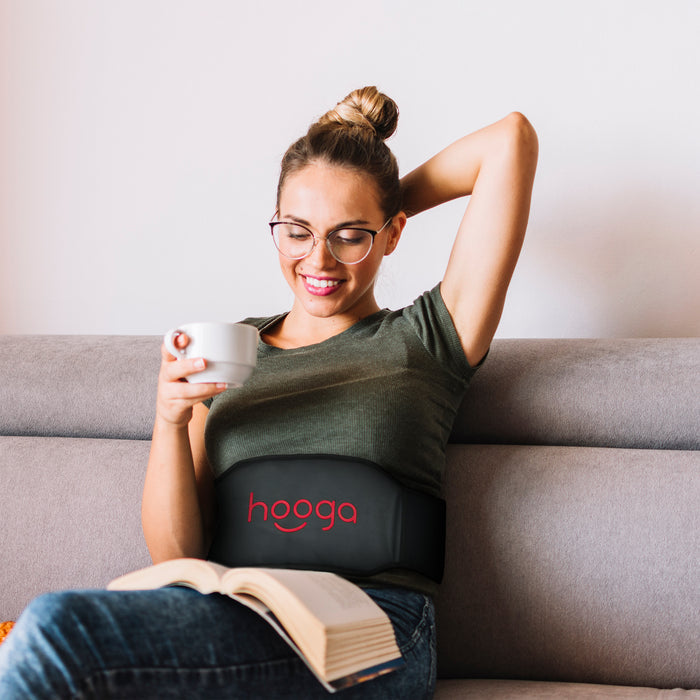 Woman relaxing on a couch reading with the Hooga PEMF Belt wrapped around her waist for hands-free abdominal therapy session