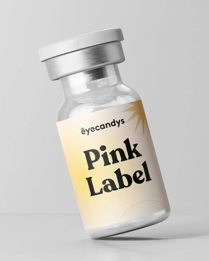 A small glass vial with a white cap labeled “EyeCandys Pink Label Angel Green” in bold black text rests on a pastel gradient background, perfect for storing natural color contact lenses. The backdrop is plain light gray.