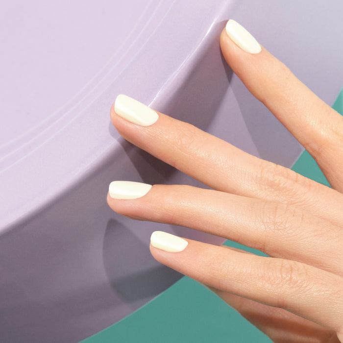 A hand with neatly manicured nails painted in Manucurist’s Porcelaine gel polish rests on the edge of a round, light purple surface against a teal background.
