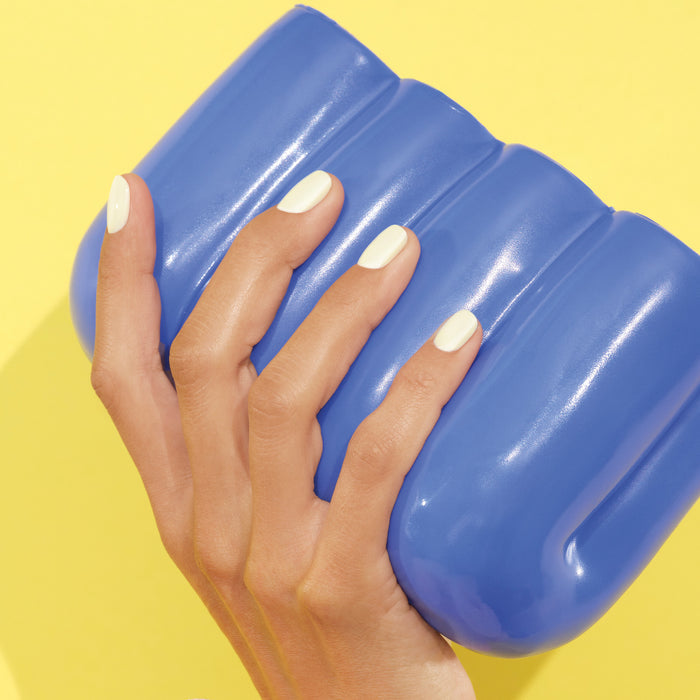 A hand with neatly manicured, off-white Porcelaine gel nails by Manucurist holds a large shiny blue object against a vivid yellow background.