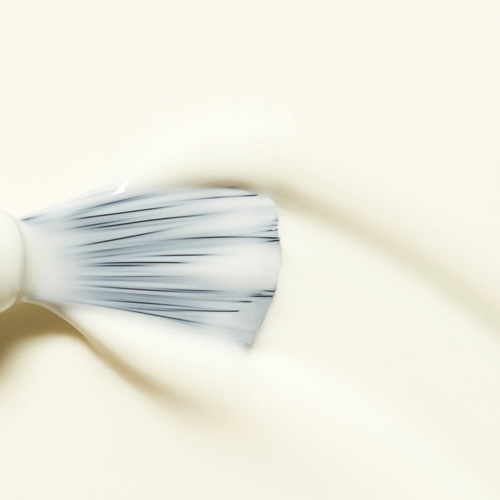 A close-up shows a brush applying Manucurist Porcelaine, a creamy off-white gel polish, onto a matching pale surface, creating soft lines and texture in the smooth layer.