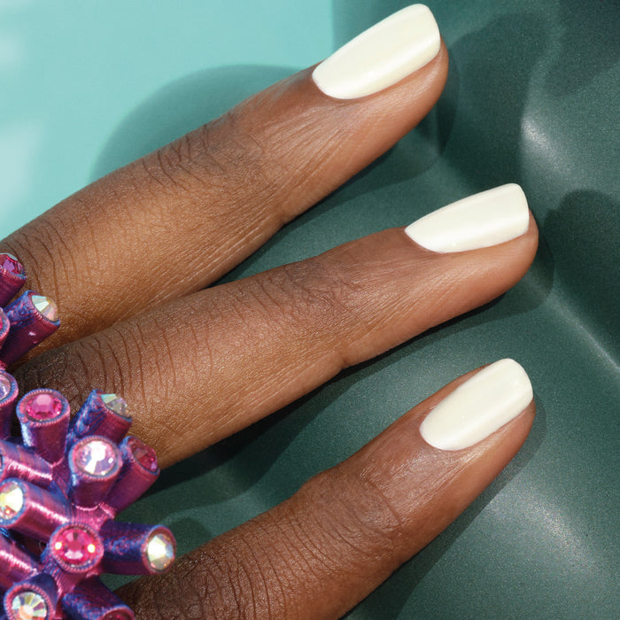 A hand with neatly manicured nails painted in Manucurist's Porcelaine gel polish and wearing a bold purple gem ring rests on a smooth green surface.
