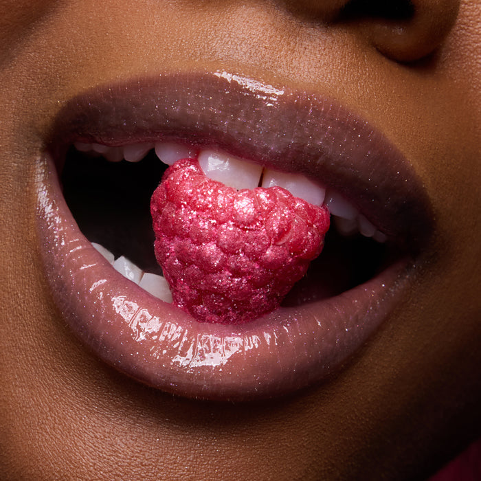 Glossy, shimmer-coated lips wearing Colourpop Fresh Kiss Glossy Balm in Raspberry Crème hold a sparkling raspberry between white teeth, promoting sweet, juicy Lip Gloss & Lip Oil shine
