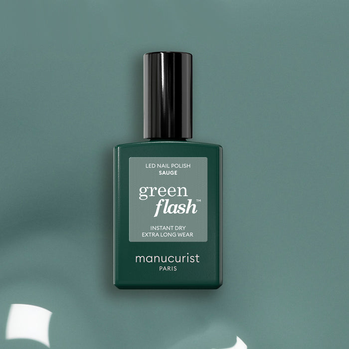 A bottle of Manucurist Sage Green Flash LED nail polish stands upright on a sage green surface, featuring a glossy black cap and minimalist label that highlights its soft green shade.