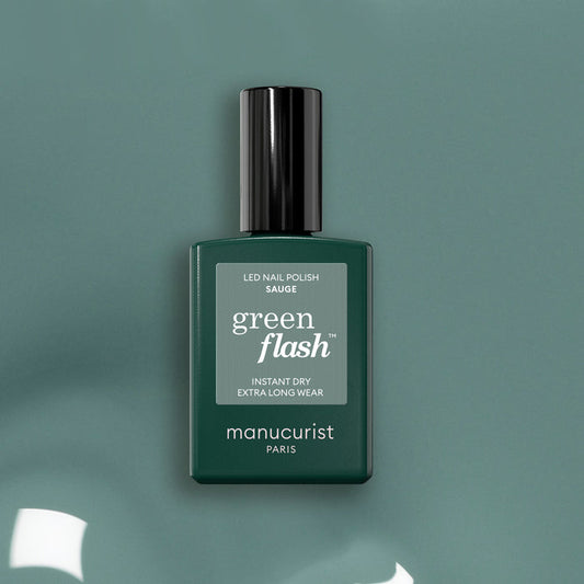 A bottle of Manucurist Sage Green Flash LED nail polish stands upright on a sage green surface, featuring a glossy black cap and minimalist label that highlights its soft green shade.