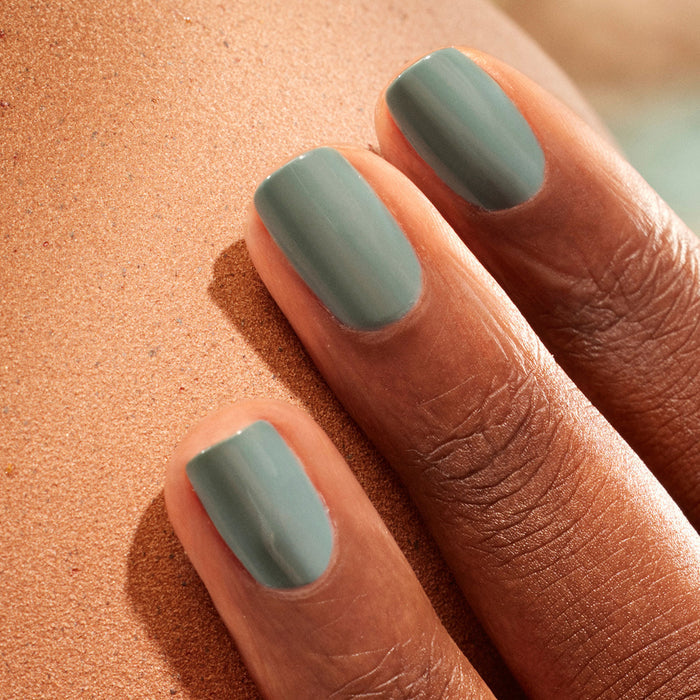 A close-up of a dark-skinned hand with neatly manicured nails painted in Manucurist's Sage, a soft green shade, resting on a textured surface.
