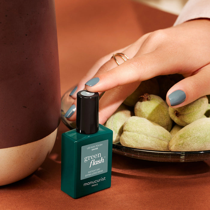 A hand with sage green-painted nails rests on a bowl of green almonds. Nearby is Manucurist Sage nail polish, matching the soft green shade, set on a brown surface.