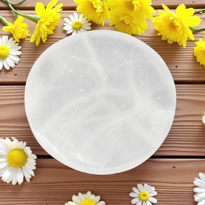 Round white selenite charging plate on a wooden surface with yellow and white flowers around it