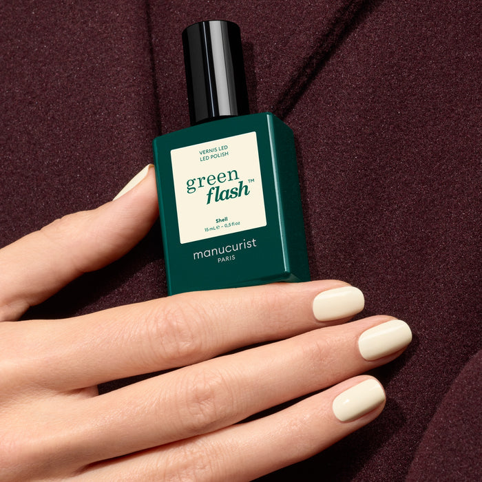 A hand with cool-toned, off-white nail polish holds a bottle of Shell by Manucurist, set against a rich burgundy background.