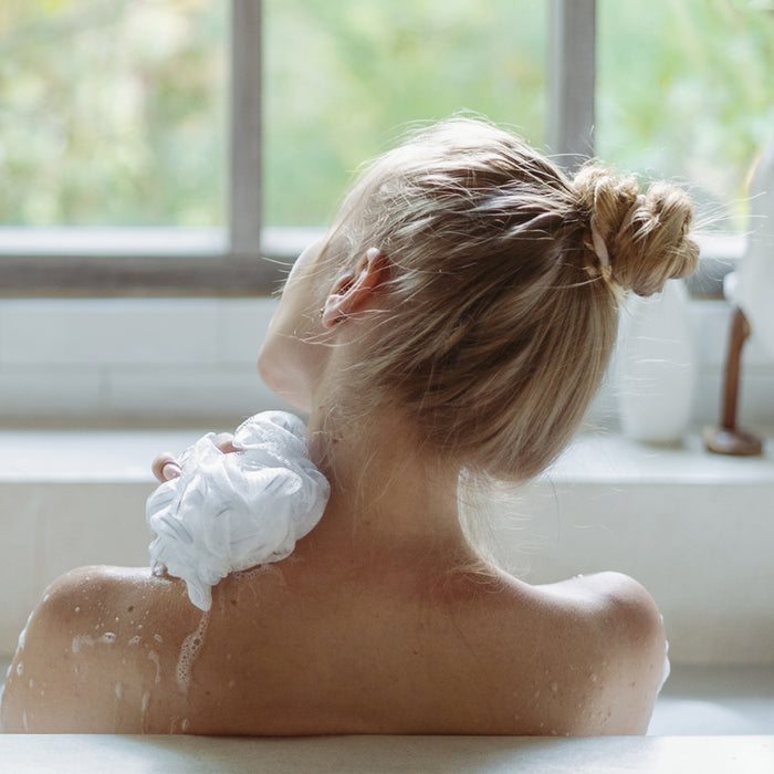 Woman's back in a bathtub washing her back with a mesh sponge and a Deluvia soap bar