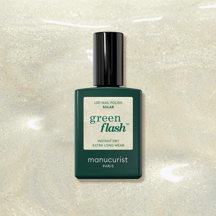 A bottle of Manucurist Solar nail polish rests on a shimmery pale beige background with hints of golden shimmer. The rectangular green bottle features a black cap and a label displaying white and green text.