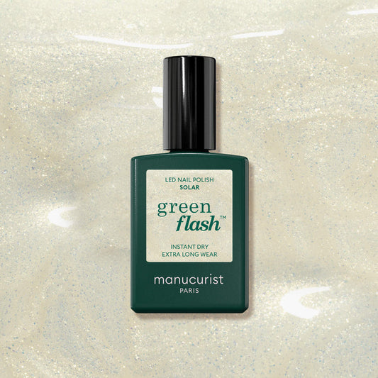 A bottle of Manucurist Solar nail polish rests on a shimmery pale beige background with hints of golden shimmer. The rectangular green bottle features a black cap and a label displaying white and green text.