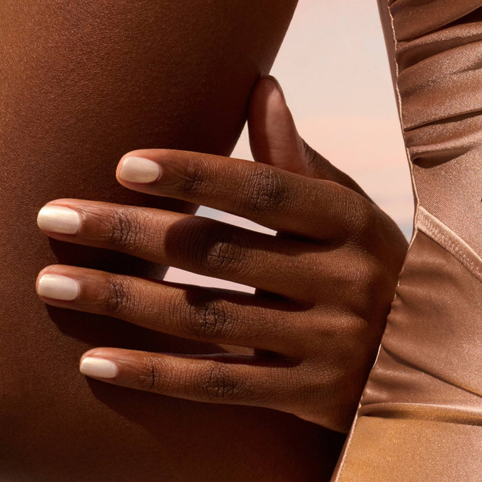 A close-up of a hand with Manucurist's Solar light beige nail polish resting on a bent knee, wearing a shimmery taupe-gold garment against a softly blurred background.