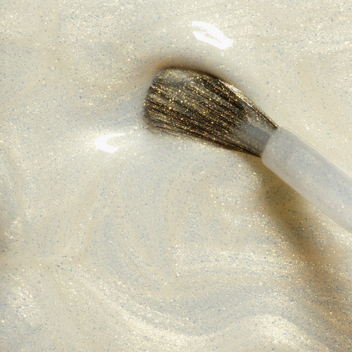 A close-up of the Manucurist Solar nail polish brush, swirling with shimmering pale gold glitter, creates a glossy, sparkly texture enhanced by golden shimmer.