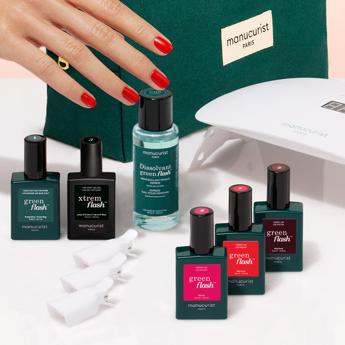 A hand with red polished nails rests on a green Manucurist bag, surrounded by the Manucurist Starter Kit, which includes nail polish, vitaminized base coat, gel polish kit, remover, UV lamp, and care accessories.