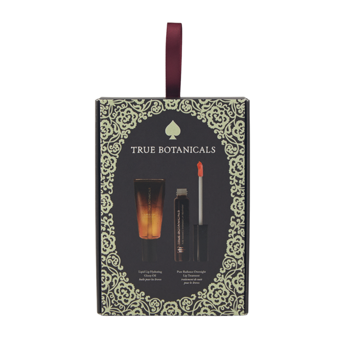 A front view of a vertical, rectangular dark green gift box from True Botanicals against a solid white background. The box is styled like a hanging holiday ornament with a loop of burgundy ribbon attached to the top. An intricate light-gold filigree pattern creates a thick decorative frame around the front edge. Inside the frame, a gold ace-of-spades-style logo sits above the brand name "TRUE BOTANICALS." The center displays graphic illustrations of the two included items: a small squeezable tube of Lipid L
