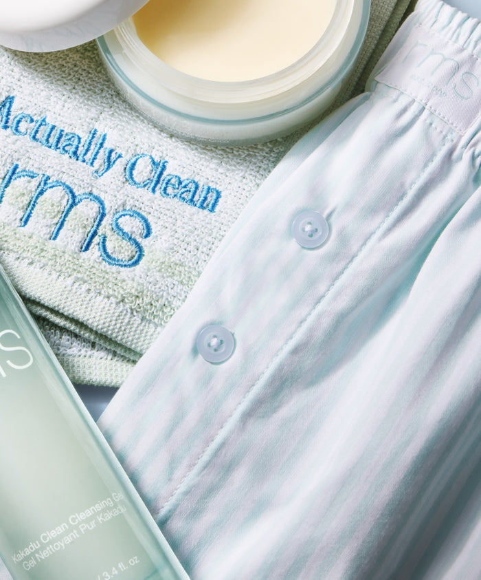 Light pastel striped Striped Sleep Shorts with buttons, placed next to skincare products and a towel embroidered with RMS, emphasizing clean beauty and natural ingredients.
