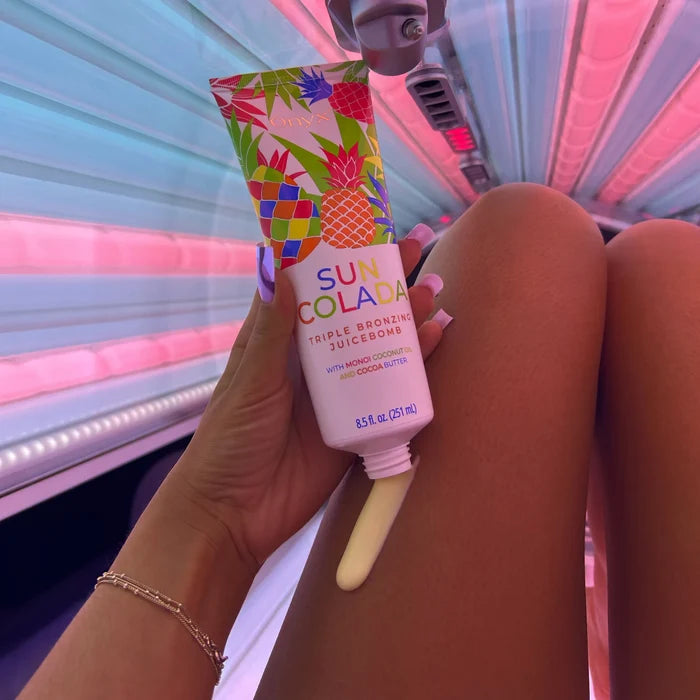 Kobieta w solarium trzyma balsam Onyx Suncolada, z tubki wyciśnięta kremowa porcja balsamu na nodze, różowo-fioletowe oświetlenie kabiny