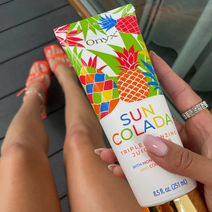Kobieta trzyma balsam Onyx Suncolada na tle ciemnej podłogi, widoczne opalone nogi i pomarańczowe sandały, produkt z motywem tropikalnych owoców