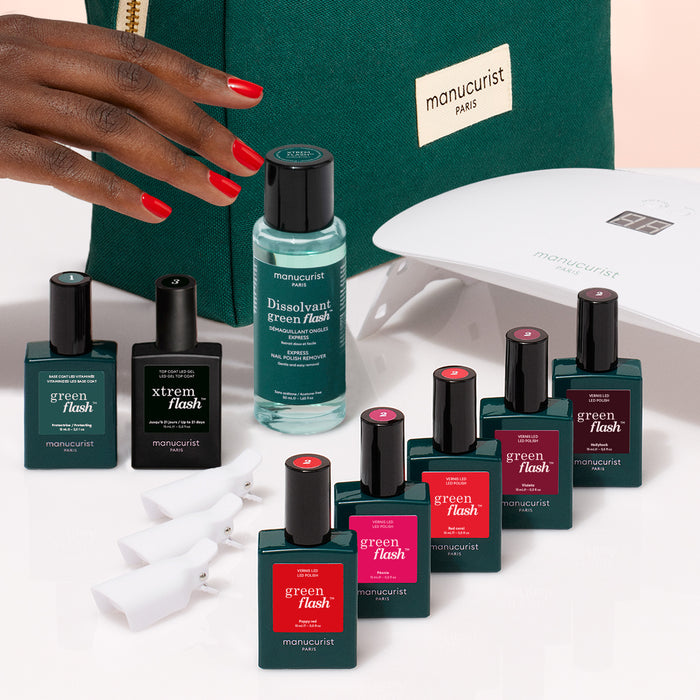 A hand with red nails reaches for a green Manucurist bag. In front are items from the Manucurist Super Starter Kit: Green Flash gel polish, remover, a black bottle, nail clips, and a white LED lamp for a lasting manicure.