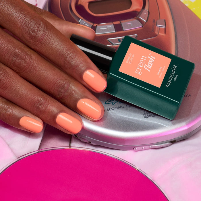 A hand with glossy, nineties-inspired nails rests on a portable CD player, next to a bottle of Manucurist's Tangerine gel polish. A partially visible pink CD sits in the foreground.