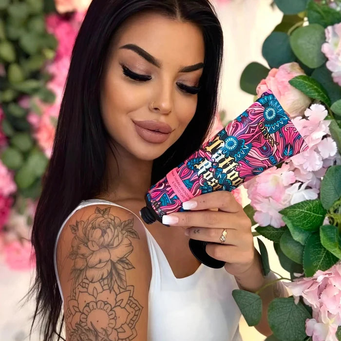 Kobieta w białym topie trzyma tubę Onyx Tattoo Rose na tle różowych kwiatów i zielonych liści.