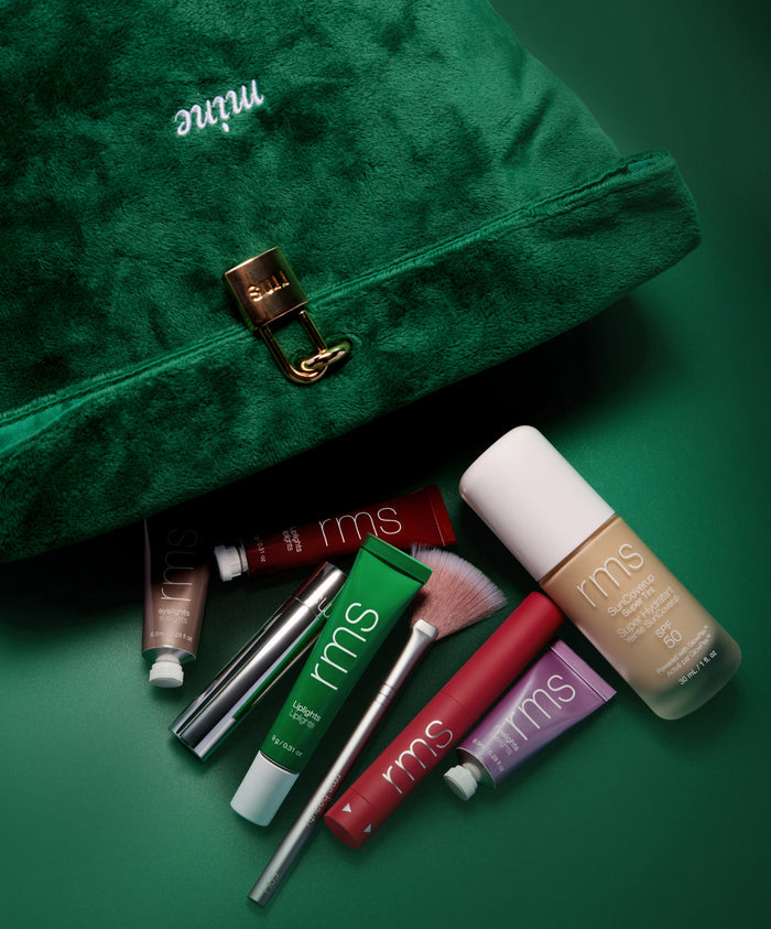The Emerald Luxe Pouch in rich green velvet with gold clasp, surrounded by RMS Beauty skincare and makeup products including foundation, lip tints, and brushes on a matching green background.