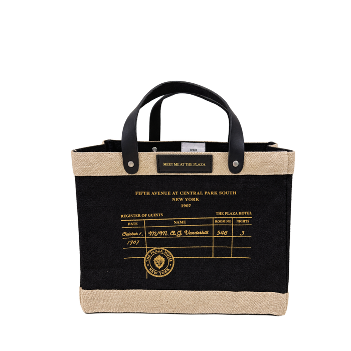 A tote bag with a black body, natural jute trim, and black leather handles. The front features a printed vintage hotel guest register design in gold with the text 'FIFTH AVENUE AT CENTRAL PARK SOUTH NEW YORK 1907' and a gold 'The Plaza Hotel New York' emblem.