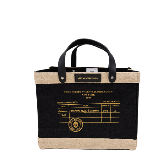 A tote bag with a black body, natural jute trim, and black leather handles. The front features a printed vintage hotel guest register design in gold with the text 'FIFTH AVENUE AT CENTRAL PARK SOUTH NEW YORK 1907' and a gold 'The Plaza Hotel New York' emblem.