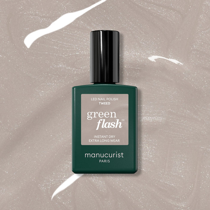 A bottle of Manucurist Tweed Green Flash LED Gel Polish rests on a shimmery beige surface, highlighting its warm grey pearlescent finish. The dark green rectangular bottle features a black cap.