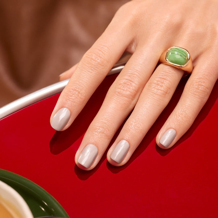 A hand with neatly manicured nails painted in Manucurist's Tweed, a soft grey shade with pearly undertones, rests on a shiny red surface. The person wears a bold gold ring set with a large green stone on their index finger.
