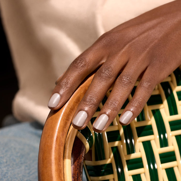A hand with neatly manicured, light beige nails polished in "Tweed" by Manucurist rests on the curved wooden arm of a woven chair with green and yellow accents. The softly blurred background adds to the elegant look.