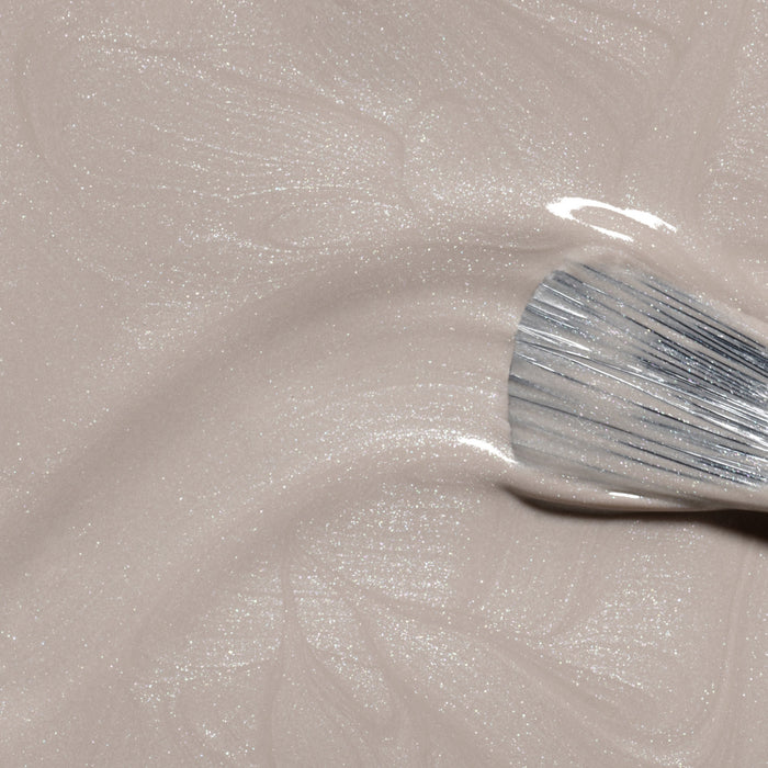 A close-up of a brush applying Manucurist Tweed, a shimmery light beige nail polish with pearly undertones, creating gentle swirls in the glossy finish.